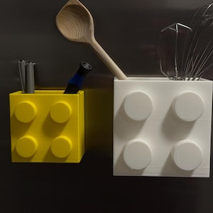 May include: Two block-shaped containers, one yellow and one white, with circular studs. The yellow container holds pens and a blue marker. The white container holds a wooden spoon, a whisk, and a spatula. Kitchen utensil holders.