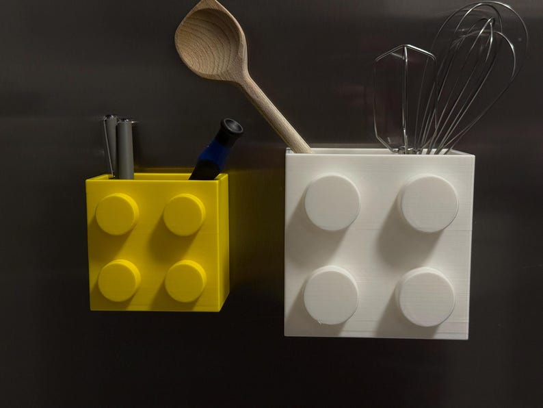 May include: Two block-shaped containers, one yellow and one white, with circular studs. The yellow container holds pens and a blue marker. The white container holds a wooden spoon, a whisk, and a spatula. Kitchen utensil holders.