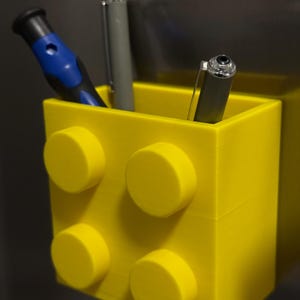 May include: A bright yellow pen holder designed to resemble a large building block. The holder contains a blue and black screwdriver and two pens. The pen holder has four yellow circular studs on the front.