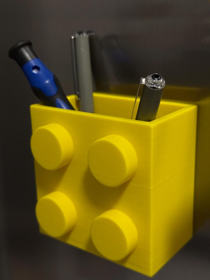 May include: A bright yellow pen holder designed to resemble a large building block. The holder contains a blue and black screwdriver and two pens. The pen holder has four yellow circular studs on the front.