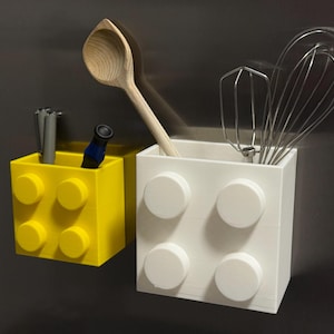 Könnte beinhalten: Zwei blockförmige Behälter, einer gelb und einer weiß, mit kreisförmigen Details. Der gelbe Behälter enthält Stifte und einen kleinen blauen Gegenstand. Der weiße Behälter enthält einen Holzlöffel und einen Schneebesen. Küchenaufbewahrung.