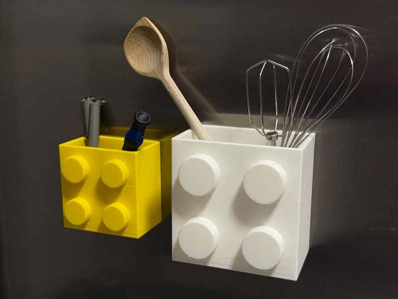 May include: Two block-shaped containers, one yellow and one white, with circular details. The yellow container holds pens and a small blue object. The white container holds a wooden spoon and a whisk. Kitchen storage.