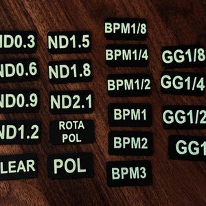 May include: A set of 14 black rectangular filter labels with white text. The labels include ND0.3, ND0.6, ND0.9, ND1.2, ND1.5, ND1.8, ND2.1, CLEAR, POL, ROTA POL, BPM1/8, BPM1/4, BPM1/2, BPM1, BPM2, BPM3, GG1/8, GG1/4, GG1/2, GG1.
