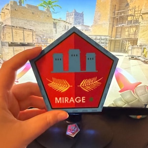 May include: A pentagon-shaped sticker with a red background and a blue border. The sticker features three blue tower illustrations at the top, two golden leaf illustrations at the bottom, and the word "MIRAGE" in white.