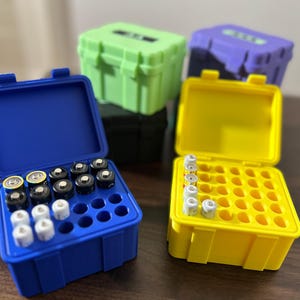 Könnte beinhalten: Vier Batteriekästen, zwei blau, einer gelb und einer grün. Die blauen Kästen sind geöffnet und zeigen Reihen von Batterien im Inneren. Der gelbe Kasten ist geöffnet und zeigt leere Batteriefächer.