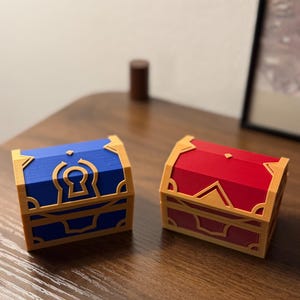 Könnte beinhalten: Zwei Miniatur-Schatzkisten, eine blaue und eine rote, mit goldfarbenen Verzierungen. Jede Truhe hat ein Schlüsselloch-Design auf der Vorderseite und dekorative Akzente. Die Truhen werden auf einer Holzoberfläche präsentiert.