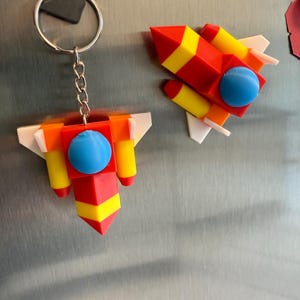 Peut inclure: Un porte-clés et un aimant en forme de fusée rouge, jaune et bleu. Le porte-clés a une chaîne et un anneau argentés. La fusée a une sphère bleue au centre, avec des accents jaunes et rouges. L'aimant est une fusée assortie.