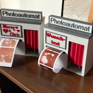 Könnte beinhalten: Zwei Miniatur-Fotoautomaten-Modelle mit dem Wort "Photoautomat" oben. Einer hat "Merve & Chris" und ein Herz, der andere "Wunschtext". Beide haben rote Vorhänge und Fotostreifen.