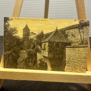 Puede incluir: Una postal antigua en tonos sepia con una vista paisajística de Rothenburg ob der Tauber, Alemania. La imagen muestra fortificaciones de piedra, una torre y un pequeño edificio con techo de tejas. El texto dice: Rothenburg o. T. Wallpartie am Rödertor.