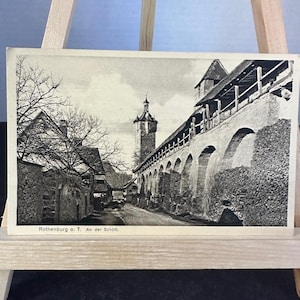 Op de afbeelding: Een zwart-wit ansichtkaart met een historische stenen muur met bogen, een hoge toren en kale bomen langs een pad. De tekst Rothenburg o. T. An der Schütt staat gedrukt in de linkerbenedenhoek van de rechthoekige papieren kaart.
