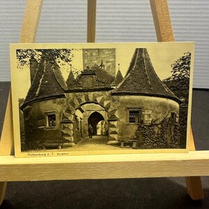 May include: A vintage sepia-toned postcard featuring the Burgthor gate in Rothenburg ob der Tauber. The image displays a stone fortification with two conical towers, a central arched entryway, and a person standing in the distance. Text reads Rothenburg o. T. Burgthor.