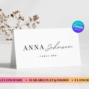 May include: White folded place card with the name "Anna Johnson" in black script. The text "- TABLE ONE -" is below the name. The card is 3.5 x 2 inches and 4 x 2 inches in size.