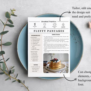 K&ouml;nnte beinhalten: Eine Rezeptkarte f&uuml;r fluffige Pfannkuchen liegt auf einem blauen Teller, mit einem Zweig Gr&uuml;n und einer Rolle wei&szlig;em Band. Die Karte enth&auml;lt eine Zutatenliste und Anweisungen sowie ein Foto von Pfannkuchen mit Beeren. Der Text auf der Karte lautet "Fluffy Pancakes".