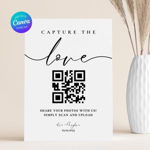 May include: A white wedding sign with the text "CAPTURE THE love" in elegant script, a QR code, and the text "SHARE YOUR PHOTOS WITH US! SIMPLY SCAN AND UPLOAD". The sign also includes a date and names. A vase with decorative stems is in the background.