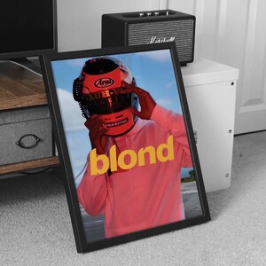 May include: A framed poster featuring a photograph of a person wearing a red racing helmet with the word "blond" in yellow lettering on a pink background.