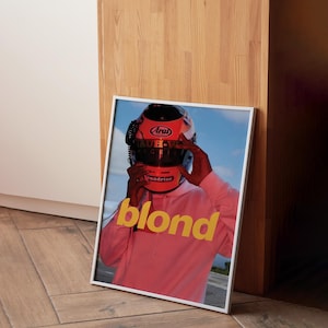May include: A framed poster featuring a person wearing a red racing helmet and a pink hoodie. The word "blond" is printed in yellow letters on the poster.