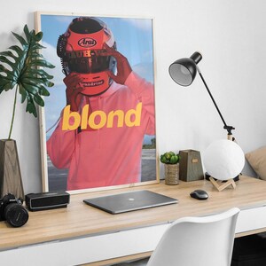 May include: A framed poster featuring a person wearing a red racing helmet with the word "blond" in yellow letters on a pink background.