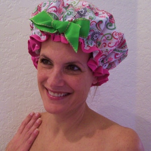 May include: A floral shower cap with a pink trim and a green bow. The cap features a paisley pattern in pink, green, and white. The bow is a bright green with pink stitching.