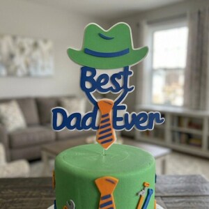 Könnte beinhalten: Ein Tortenstecker mit einem grünen Fedora-Hut über dem blauen Schriftzug "Best Dad Ever" und einer orangefarbenen Krawatte mit blauen Streifen. Der Stecker steckt in einer grün glasierten Torte, die mit kleinen orangefarbenen Krawatten und blauen Werkzeugsymbolen verziert ist.