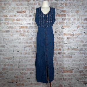 May include: A sleeveless, button-down denim dress with floral embroidery on the bodice. The dress is a medium blue color and has a V-neckline. The dress is displayed on a mannequin against a brick wall.