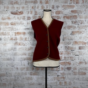May include: A burgundy sleeveless vest with a brown trim and button closure. The vest is displayed on a white mannequin against a white brick wall.