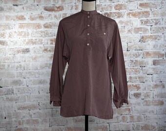 May include: A long-sleeved, brown tunic-style shirt with a mandarin collar and a button-down placket. The shirt has a single chest pocket and is displayed on a black mannequin against a brick wall background.