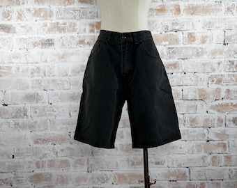 May include: Black denim shorts displayed on a mannequin. The shorts have a high waist, a button and zipper closure, and pockets. The shorts are knee-length and have a relaxed fit. The mannequin is set against a white brick wall.