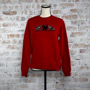 May include: Red sweatshirt with a crew neck and long sleeves. The sweatshirt features an embroidered design of three cats, two with leopard print and one black cat.