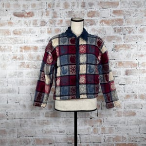 May include: A red, blue, and cream patterned fleece jacket with a zipper closure. The jacket has a patchwork design with various motifs, including birds, hearts, and apples.