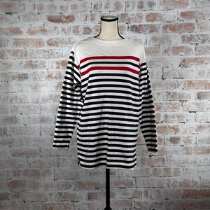 May include: A long-sleeved shirt with a white upper section and navy and white horizontal stripes. A red stripe accents the chest area. The shirt is displayed on a mannequin against a brick wall background.