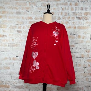 May include: A bright red long-sleeve cardigan featuring a button-down front and ribbed cuffs. The garment is decorated with embroidered pink heart motifs and swirling vine patterns on the front panels, creating a distinct visual contrast.