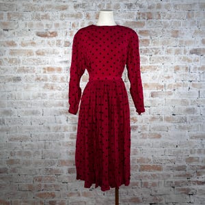 May include: A red dress with black polka dots. The dress has long sleeves and a midi-length skirt. The dress is displayed on a mannequin against a brick wall background. The dress has a fitted waist and a round neckline.