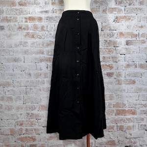 May include: A long, black skirt with a button-down front. The skirt is made of a flowing fabric and has a high waist. The skirt is displayed on a mannequin against a brick wall background.