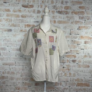 May include: Cream-colored button-down shirt with short sleeves. The shirt features embroidered patches in various colors and patterns, including floral and plaid designs. The shirt is displayed on a mannequin against a brick wall background.