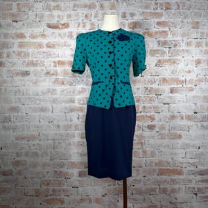 May include: A teal and navy blue two-piece suit displayed on a mannequin. The jacket has black polka dots and black buttons. The skirt is a solid navy blue. The suit is a vintage style.