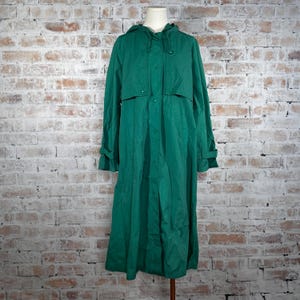 May include: A long, emerald green raincoat with a hood, button closures, and long sleeves. The coat is displayed on a mannequin against a brick wall background. The coat has a classic, vintage style.