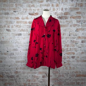 May include: A red button-down shirt with long sleeves and a collar. The shirt features a black floral pattern. The shirt is displayed on a mannequin against a brick wall background.
