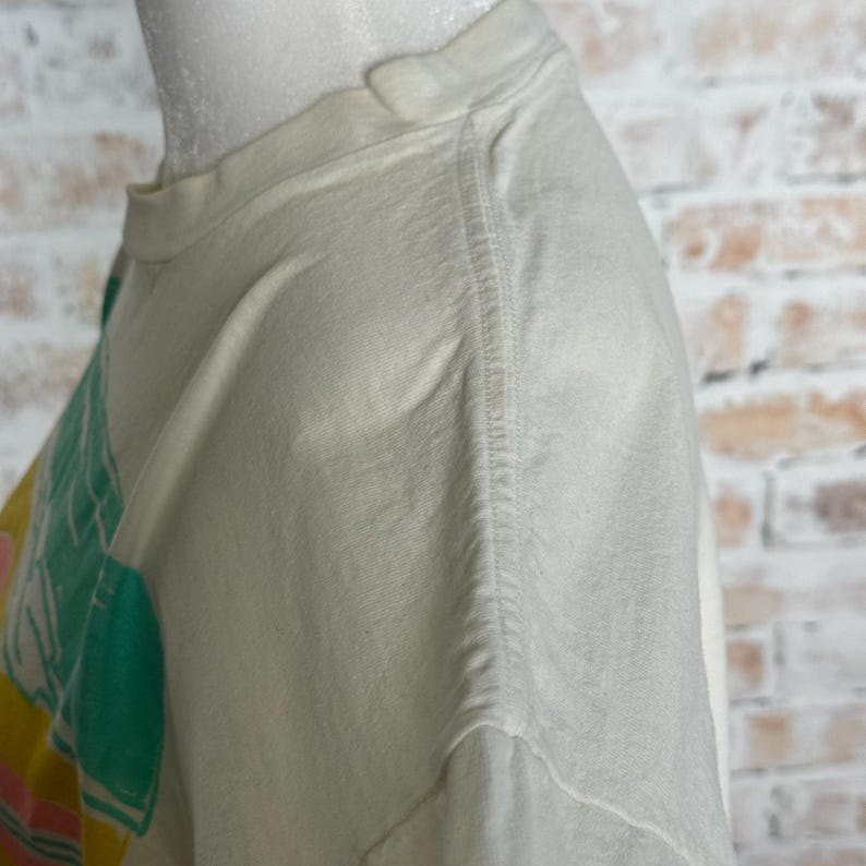 This image showcases the right shoulder of a true vintage 1980s salvage condition thrashed Belton Tough Tee women’s 100% preshrunk cotton St. Thomas single stitch graphic t-shirt in a size XL modeled on a dress form in front of a brick backdrop.