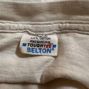 This image showcases a close up view of the interior tag on a true vintage 1980s salvage condition thrashed women’s 100% preshrunk cotton St. Thomas single stitch graphic t-shirt in a size XL. The tag indicates that the top was made by Belton Tough Tee.