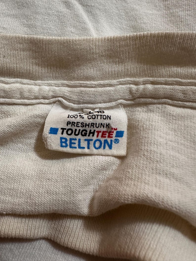 This image showcases a close up view of the interior tag on a true vintage 1980s salvage condition thrashed women’s 100% preshrunk cotton St. Thomas single stitch graphic t-shirt in a size XL. The tag indicates that the top was made by Belton Tough Tee.