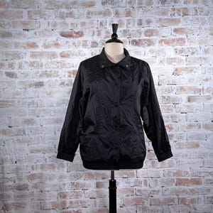 May include: A black jacket with a collared neckline and button-down front. The jacket has long sleeves with ribbed cuffs and two front pockets. It is displayed on a mannequin against a brick wall background.