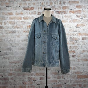 May include: A light blue denim jacket with a classic design. The jacket features a button-up front, two chest pockets, and long sleeves. It is displayed on a mannequin against a brick wall background.