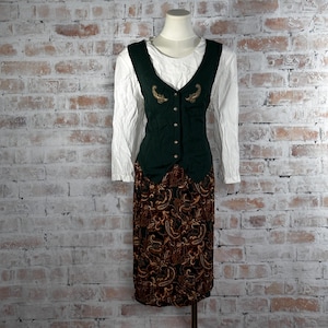 May include: A vintage ensemble featuring a white long-sleeve shirt, a dark green vest with gold embroidery, and a paisley print skirt. The vest has a button-down closure. The skirt is a midi length.