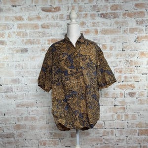 May include: A short-sleeved button-down shirt with a brown and blue floral pattern. The shirt is displayed on a mannequin against a brick wall background. The fabric has a detailed, ornate design.