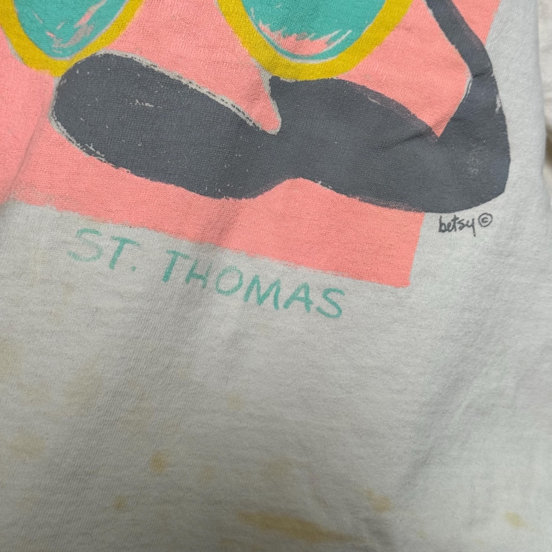 This image showcases a close up view of the logo below the print on the front side of a true vintage 1980s salvage condition thrashed Belton Tough Tee women’s 100% preshrunk cotton St. Thomas single stitch graphic t-shirt in a size XL modeled on a dress form.