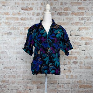 May include: A short-sleeve button-down shirt featuring a bold, abstract floral pattern in vibrant shades of teal, royal blue, and purple set against a solid black background. The garment has a classic collar and a relaxed, loose-fitting silhouette.