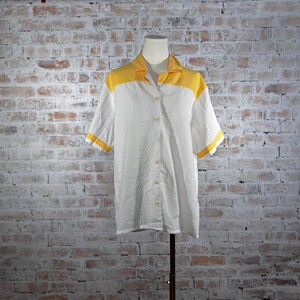 May include: White button-down shirt with short sleeves and a yellow collar and sleeve trim. The shirt is displayed on a mannequin against a brick wall background. The shirt has yellow buttons.