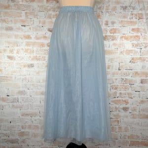 May include: A light blue, floor-length skirt made of a sheer, flowing fabric. The skirt has an elastic waistband and a soft, delicate appearance. The skirt is displayed against a brick wall background.
