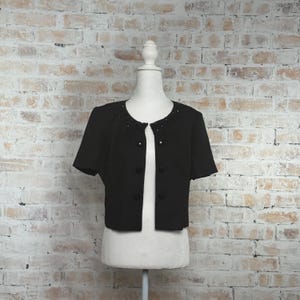 May include: A black short-sleeved bolero jacket displayed on a mannequin. The jacket has a round neckline embellished with small, dark beads and features three buttons down the front. The garment is made of a smooth, dark fabric.