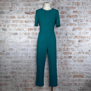 May include: Teal green jumpsuit with short sleeves and a round neckline. The one-piece garment has a textured pattern and a fitted waist. The pants are straight-legged. The jumpsuit is displayed on a mannequin against a brick wall background.